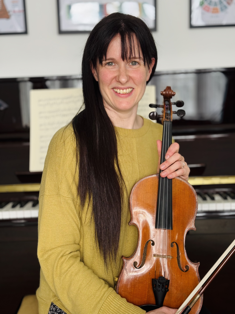 Rosie is sat on a piano stool, facing away from the piano, holding a violin in her lap.