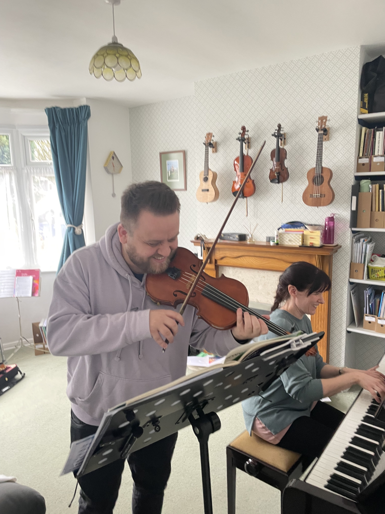 A student stand playing his violin, while Rosie sits at the piano.