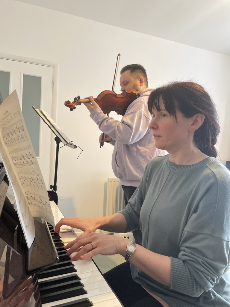 Rosie sits at her piano, accompanying her student who is standing and playing the violin
