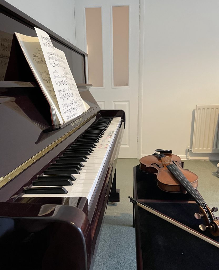 A side profile of an upright piano and stool. A viola rests on the stool.