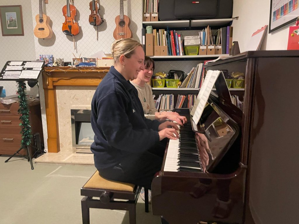 A students sits playing the piano, while Rosie sits next to them to supervise.